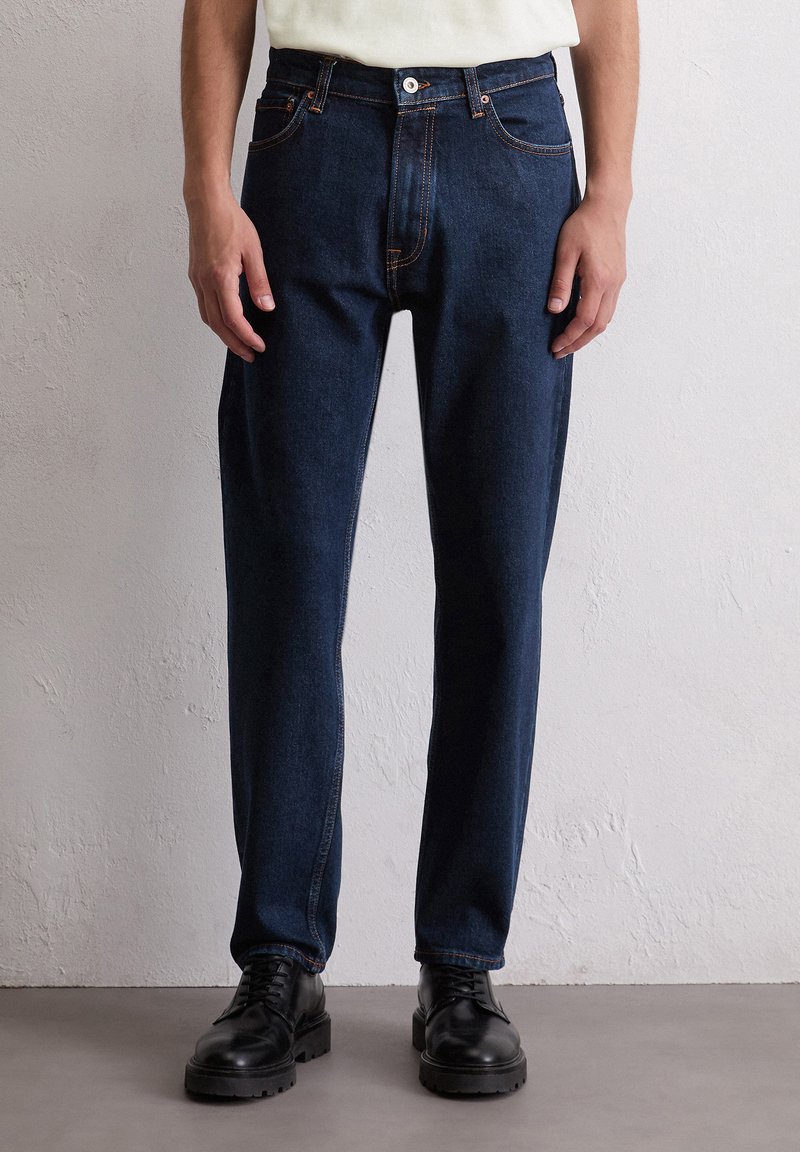 Dunkelblaue Jeans mit geradem Schnitt, ausgestattet mit orangefarbenen Nähten und einem Knopfverschluss. Kombiniert mit schwarzen Schnürstiefeln.