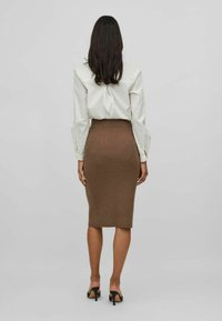 Camisa blanca de manga larga y falda midi marrón de canalé, diseño ajustado, textura suave, combinados con tacones abiertos negros, vistos desde atrás.