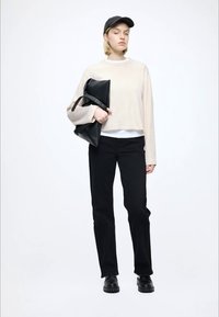 Light beige cropped sweater over a white shirt, black pants, and black footwear. Holding a black clutch bag and wearing a black cap.