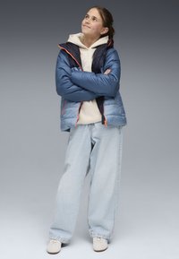 Blauer puffy Mantel mit marineblauen Akzenten, orangen Reißverschlüssen, hellbeige Kapuzenpullover und weiten hellblauen Jeans. Weiße Schuhe runden das Outfit ab.