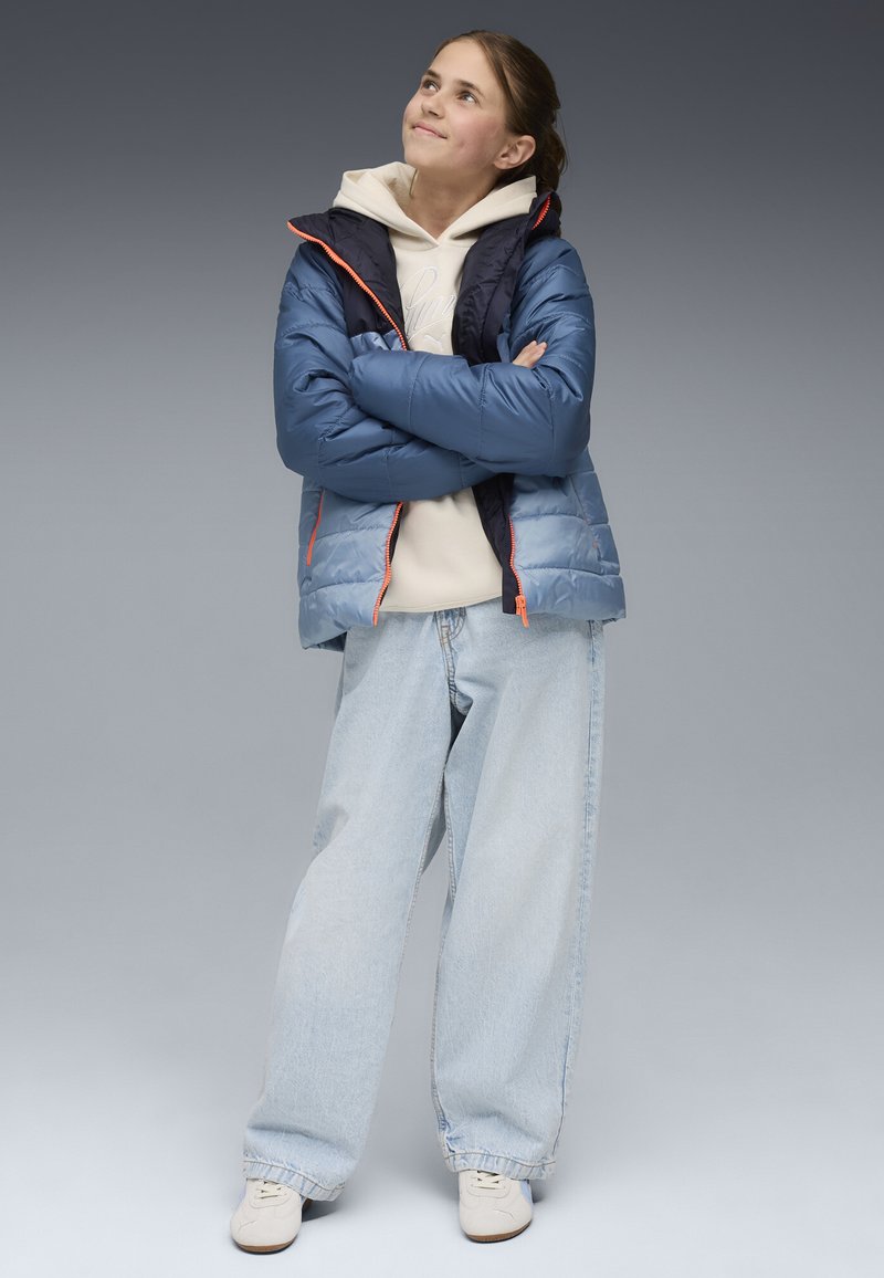 Blauer puffy Mantel mit marineblauen Akzenten, orangen Reißverschlüssen, hellbeige Kapuzenpullover und weiten hellblauen Jeans. Weiße Schuhe runden das Outfit ab.
