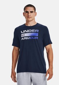 Mørkeblå kortermet t-skjorte laget av mykt stoff med "UNDER ARMOUR"-logoen i gradient hvit og blå over brystet. Enkel design.