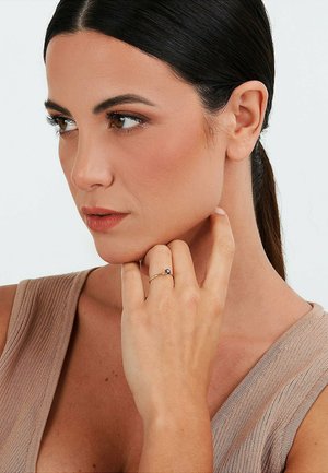 Bague en or avec une petite pierre précieuse sombre, portée au doigt. La main du modèle est positionnée près de son visage, vêtue d'un haut beige texturé.