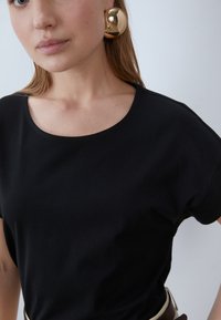 Svart kortärmad T-shirt med rund halsringning, tillverkad av slät tyg. Har stora, reflekterande guldörhängen och en minimalistisk design.