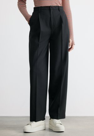 Άτομο που φοράει μαύρο παντελόνι με φαρδιά πόδια και ψηλή μέση, λευκά πλατφόρμα αθλητικά παπούτσια και μακρυμάνικη ριγέ μπλούζα με τα χέρια στις τσέπες.