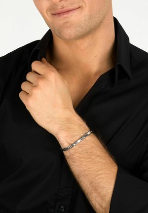 Uomo con camicia nera che sistema il colletto, indossa un braccialetto a maglie d'argento al polso sinistro su sfondo bianco.