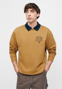 Νεαρός με σγουρά μαλλιά, φοράει μουσταρδί φούτερ με μπλε σκούρο γιακά και λογότυπο "New Balance", με τα χέρια στις τσέπες, κοιτάζει ελαφρώς προς τα δεξιά.