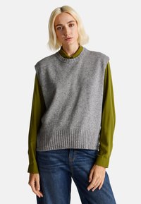 Šedý pletený svetr bez rukávů s žebrovaným lemem, nošený přes dlouhý rukáv olivově zelené blůzy. Modelka má na sobě modré džíny.