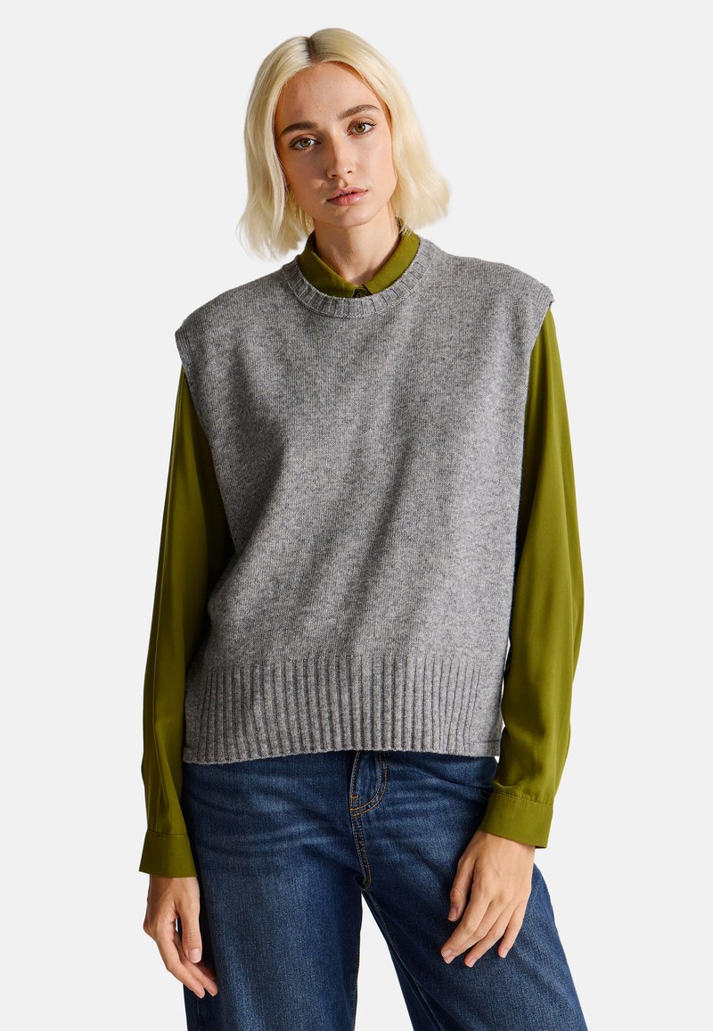 Šedý pletený svetr bez rukávů s žebrovaným lemem, nošený přes dlouhý rukáv olivově zelené blůzy. Modelka má na sobě modré džíny.