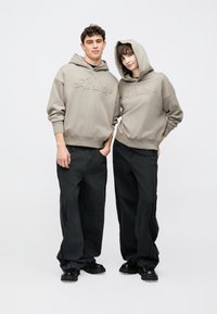 Két személy bézs színű, "Atelier" hímzéssel díszített, azonos kapucnis pulóvert visel. Mindketten fekete, bő méretű farmert és masszív fekete cipőt hordanak.