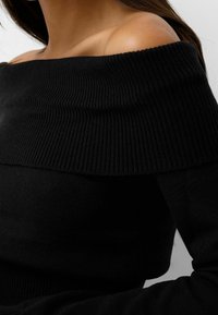 Suéter negro de canalé, con los hombros al descubierto, de textura suave, con un amplio cuello doblado y mangas ajustadas. Enfoque en los detalles del tejido.