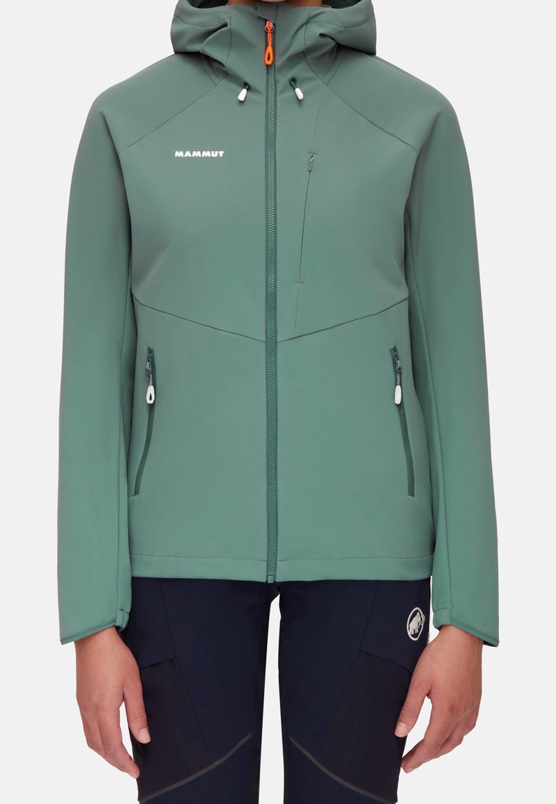 Veste softshell à capuche verte avec fermeture éclair à l'avant et poches zippées, portée avec un pantalon noir arborant un logo circulaire sur la cuisse.