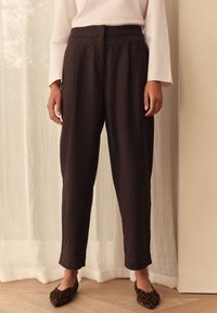 Braune, maßgeschneiderte, hoch taillierte Hosen mit Bügelfalten, zulaufenden Beinen und glatter Textur. Kombiniert mit gemusterten Loafers. Heller Hintergrund.