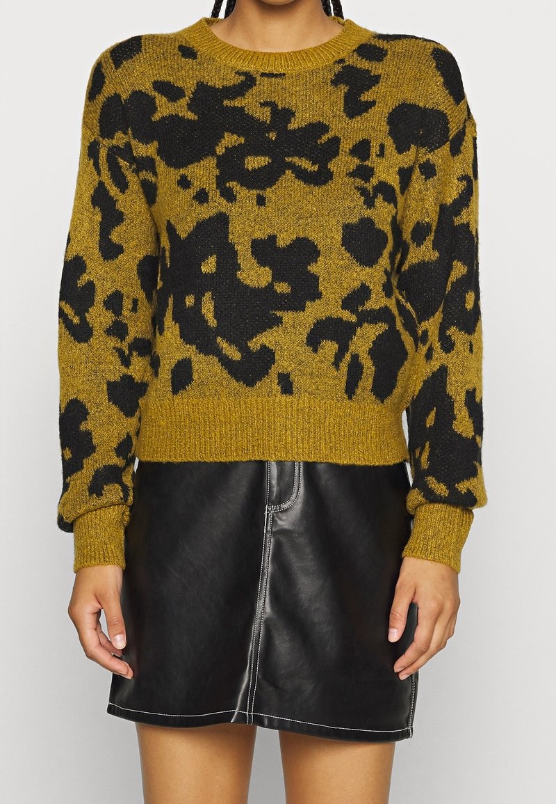Femme portant un pull à motif floral jaune moutarde et noir à manches longues et une mini-jupe en cuir noire avec des surpiqûres blanches.