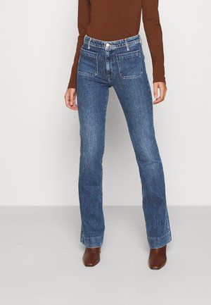 Kobieta w niebieskich dżinsach z wysokim stanem i przednimi kieszeniami naszywanymi, brązowej bluzce z długim rękawem oraz brązowych botkach, stojąca na szarej podłodze.