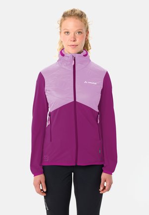 Jonge vrouw die een paars- en lavendelkleurige Vaude jas met rits draagt, staand met het gezicht naar voren en ontspannen handen langs haar zij.