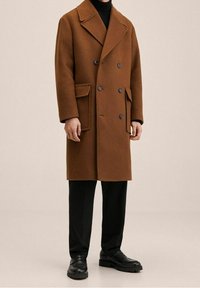 Manteau croisé en mélange de laine marron avec grands revers, poches latérales et boutons noirs, associé à un pantalon noir et des chaussures noires.