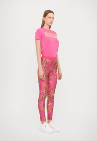 T-shirt rosa con il testo "VERSACE JEANS COUTURE", abbinato a leggings rosa con motivi floreali gialli. Sneakers bianche completano l'outfit.