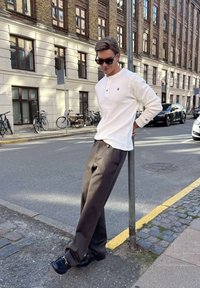 Ung mand med solbriller lænende sig mod en gadepæl på en byfortov, iført en hvid skjorte, brune bukser og sorte sko.