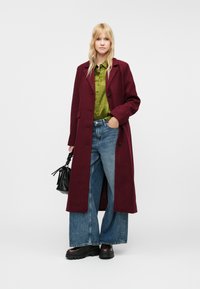 Casaco longo bordeaux sobre uma camisa verde, jeans azul de perna larga e sapatos pretos robustos. Bolsa preta com alças franjadas.