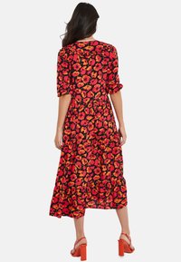 Robe longue avec un fond noir orné d'un motif floral rouge et orange, manches courtes bouffantes et ourlet à volants. Associée à des sandales rouges à talons blocs.