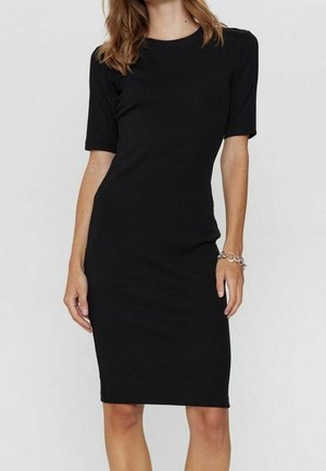 Femme portant une robe noire simple longueur genou avec manches courtes et un bracelet argenté au poignet droit sur un fond uni.