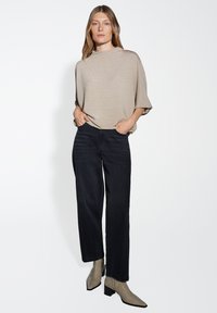 Una donna è in piedi indossando un top beige largo, jeans neri a gamba larga e stivaletti grigi sopra alla caviglia contro uno sfondo bianco uniforme.