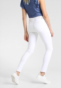 Jeans bianchi skinny con una texture liscia, vita media e dettagli sottili sulle tasche posteriori, abbinati a una camicia blu e sneakers bianche.