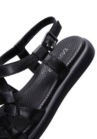 Black sandal with a perforated design, adjustable ankle strap, and a flat sole. Made of textured material with crisscross straps.