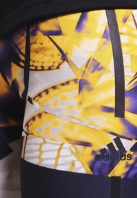 Legginsy Adidas w intensywnym żółto-fioletowym abstrakcyjnym wzorze, wykonane z gładkiego, elastycznego materiału i z czarnym pasem w talii.