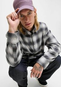 Camisa de franela a cuadros en gris y blanco con puños abotonados, combinada con jeans de mezclilla oscuros. La persona lleva una gorra de color púrpura claro con texto.