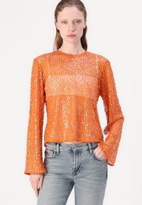Oranje top met pailletten en lange mouwen, met een ronde halslijn, featuring klokmouwen en een cropped ontwerp. Gecombineerd met blauwe jeans.