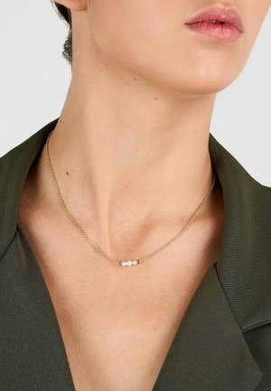 Zarte goldene Halskette mit drei kleinen weißen Perlen, getragen am Hals einer Frau, kombiniert mit einem dunkelgrünen Blazer.