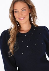 Maglione blu navy con colletto a girocollo, caratterizzato da una texture morbida e dettagli di strass sparsi. Le maniche a sbuffo aggiungono volume al design.