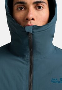 Giacca isolante blu con colletto alto, chiusura frontale con zip completa e cappuccio; presenta il logo Jack Wolfskin sul petto. Tessuto liscio e leggero.