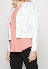 White zip-up hoodie with light purple tie-dye pattern, elastic cuffs, and waistband, worn over a pink t-shirt and black leggings.