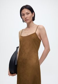 Γυναίκα με κοντά σκούρα μαλλιά, φοράει καφέ φόρεμα χωρίς μανίκια, ασημένιο κολιέ και κρατάει μια μαύρη τσάντα σε ένα απλό φόντο.