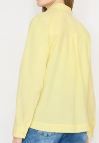 Femme portant une chemise jaune clair à manches longues et un jean bleu, vue de dos sur un fond blanc uni.