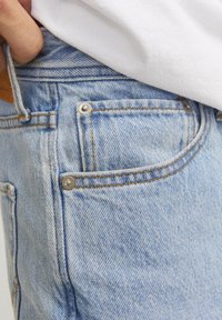 Mano con il pollice infilato nel passante della cintura di jeans di denim azzurro chiaro, indossati con una camicia bianca.