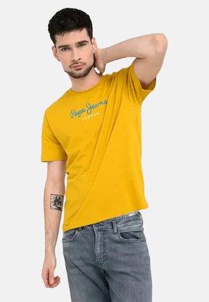 Uomo con capelli scuri che indossa una maglietta gialla Pepe Jeans e jeans grigi, posando con una mano dietro il collo e un tatuaggio sull'avambraccio sinistro.