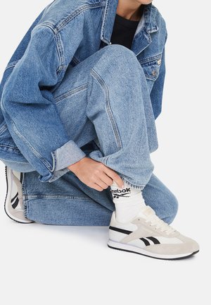 Bežové semišové tenisky Reebok s čiernymi akcentmi, obuté s bielymi ponožkami a denimovou bundou cez svetlo modré roztrhané džínsy.