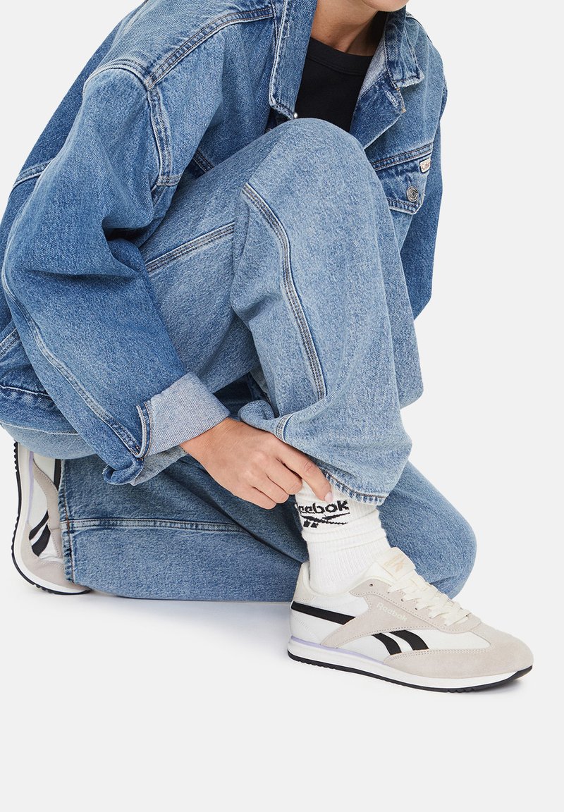 Bežové semišové tenisky Reebok s čiernymi akcentmi, obuté s bielymi ponožkami a denimovou bundou cez svetlo modré roztrhané džínsy.