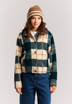Femme portant un bonnet en tricot rayé marron, une veste polaire à boutons à carreaux crème et verts, et un jean bleu, debout devant un fond clair uni.