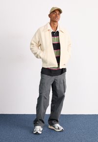Giacca in pile color crema con colletto, indossata sopra un polo a righe nere e verdi. Pantaloni cargo grigi e cappellino beige completano l'outfit.