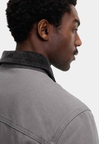 Grå stofjakke med en tekstureret sort fløjlskrave, der har et struktureret design og synlige syninger.