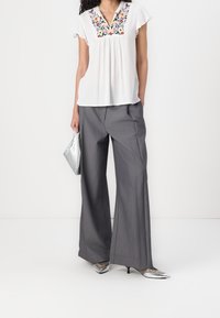 Blouse blanche avec broderies florales, pantalon large gris, pochette argentée et talons argentés pointus. Tissu lisse, coupe décontractée.