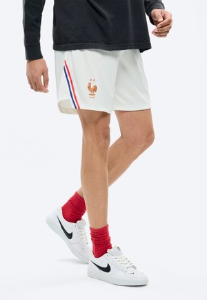 Mężczyzna ubrany w białe sportowe szorty z logo Francuskiej Federacji Piłkarskiej, czerwone skarpetki, białe buty Nike oraz czarną bluzę z długimi rękawami.