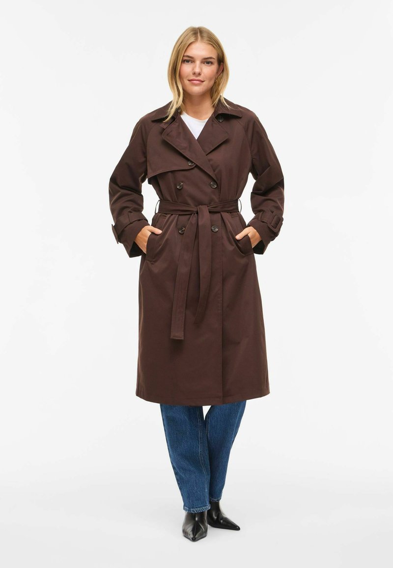Trench-coat marron croisé avec ceinture, doté de larges revers, poches latérales et coupe jusqu'aux genoux. Porté avec un jean bleu et des chaussures noires.