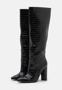 Bottes noires montant jusqu'au genou avec une finition brillante effet crocodile, bout pointu et talon bloc pour plus de stabilité.