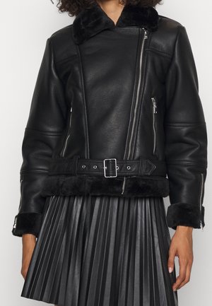 Schwarze Bikerjacke aus Kunstleder mit asymmetrischem Reißverschluss, tailliertem Gürtel, Fellkragen und Manschetten, kombiniert mit einem plissierten schwarzen Rock.